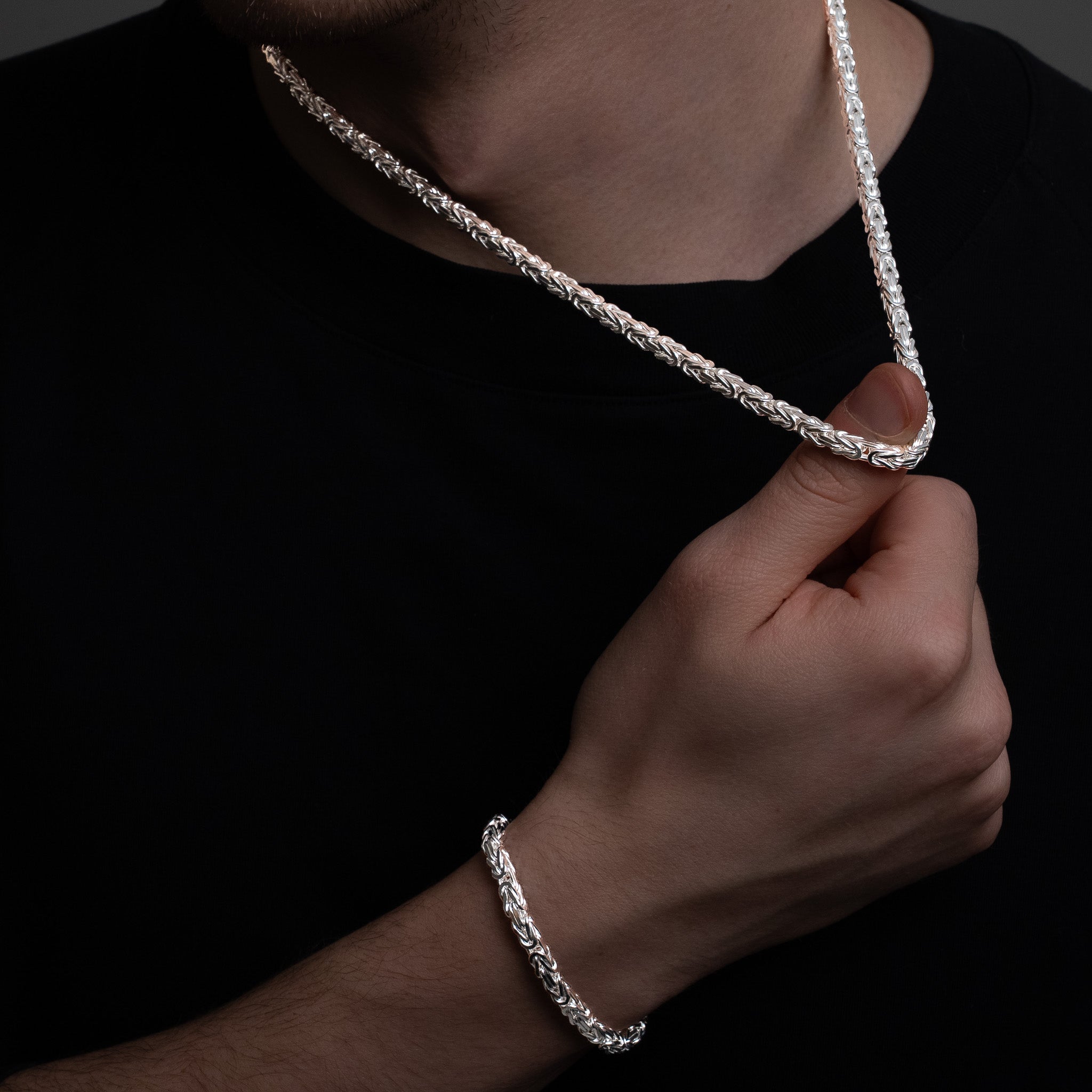 Person wearing a silver byzantine chain necklace and bracelet against a dark background