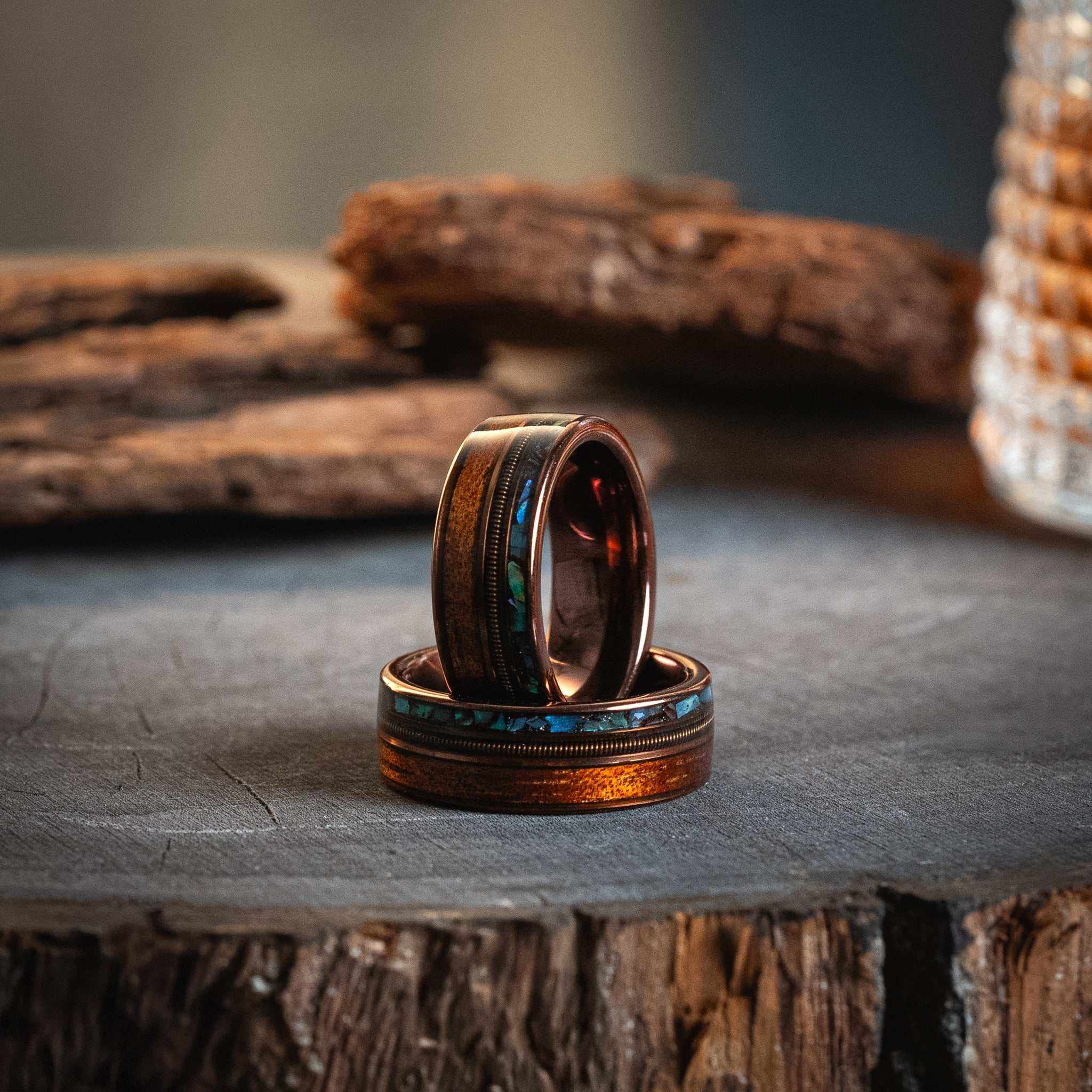 Rose gold tungsten ring with guitar string, abalone shell, and Hawaiian Koa wood inlay on rustic surface.